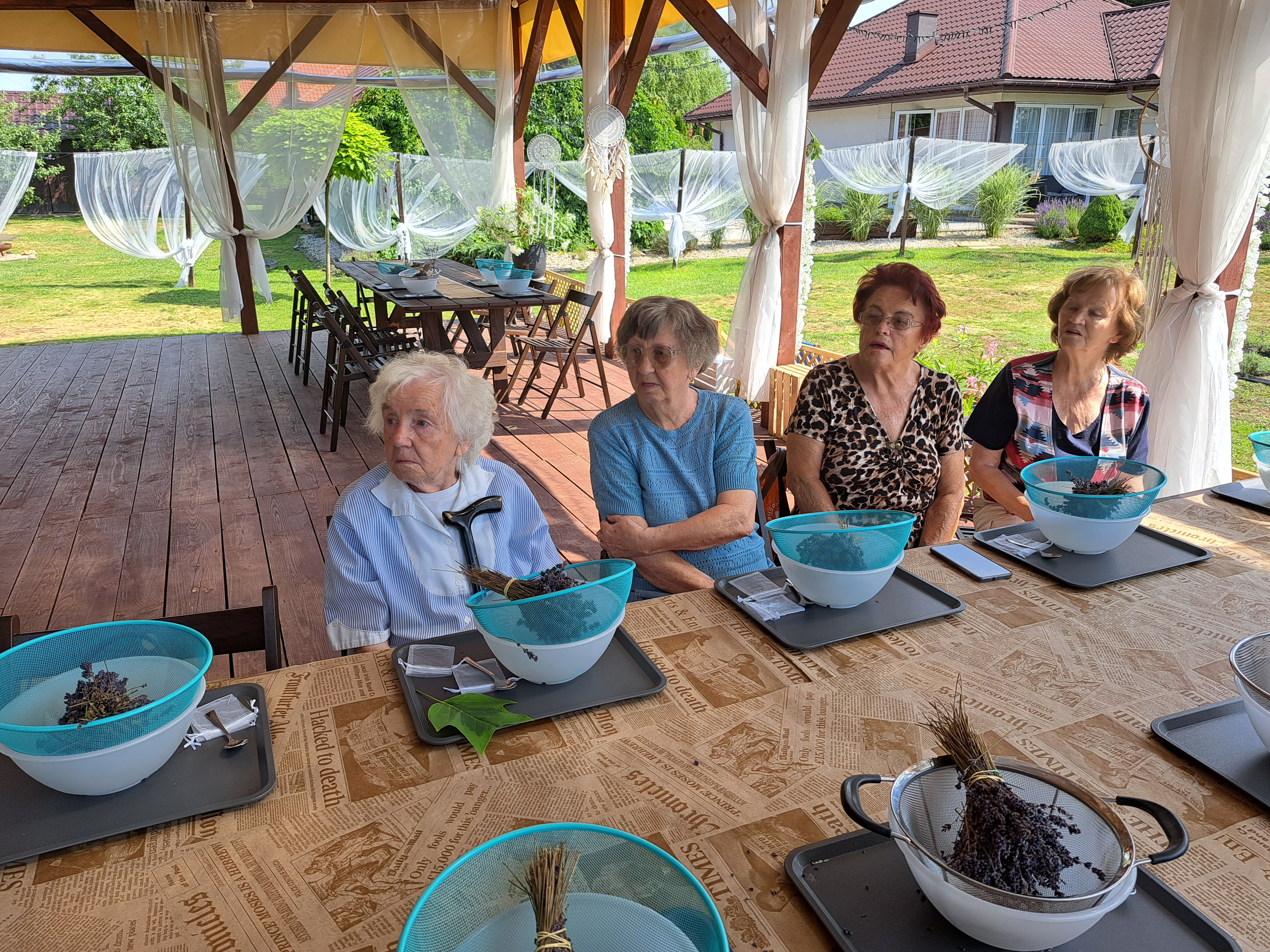 Seniorki siedzą przy stole, przed nimi stoją miski z sitkami, a w ich bukiet lawendy